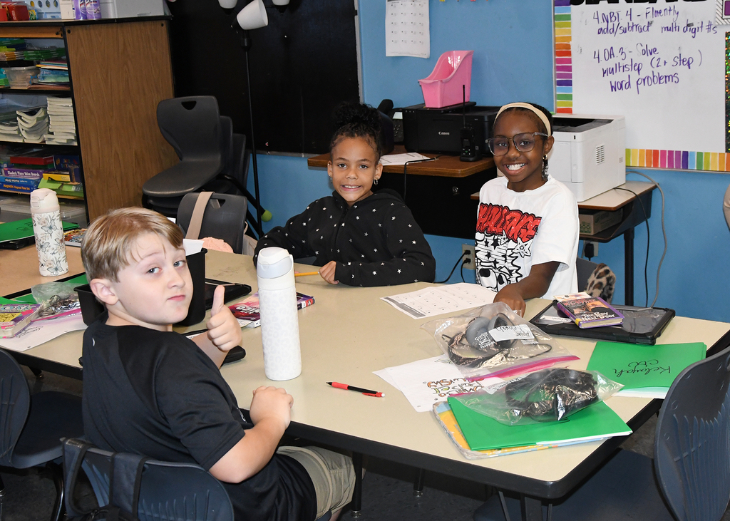 Three children seated at a desk, one giving a thumbs up. Papers, pens, and other supplies are on the desk.