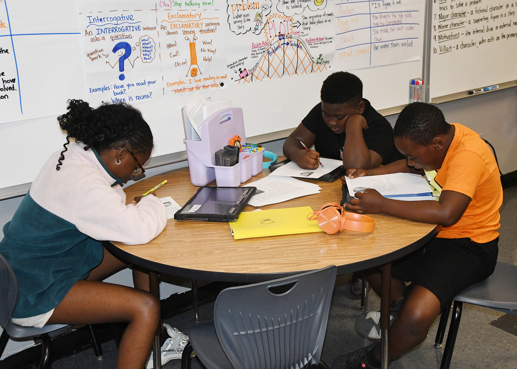 Three students sit around a round table, each with papers, a tablet, and a cup. Behind them is a whiteboard with notes.