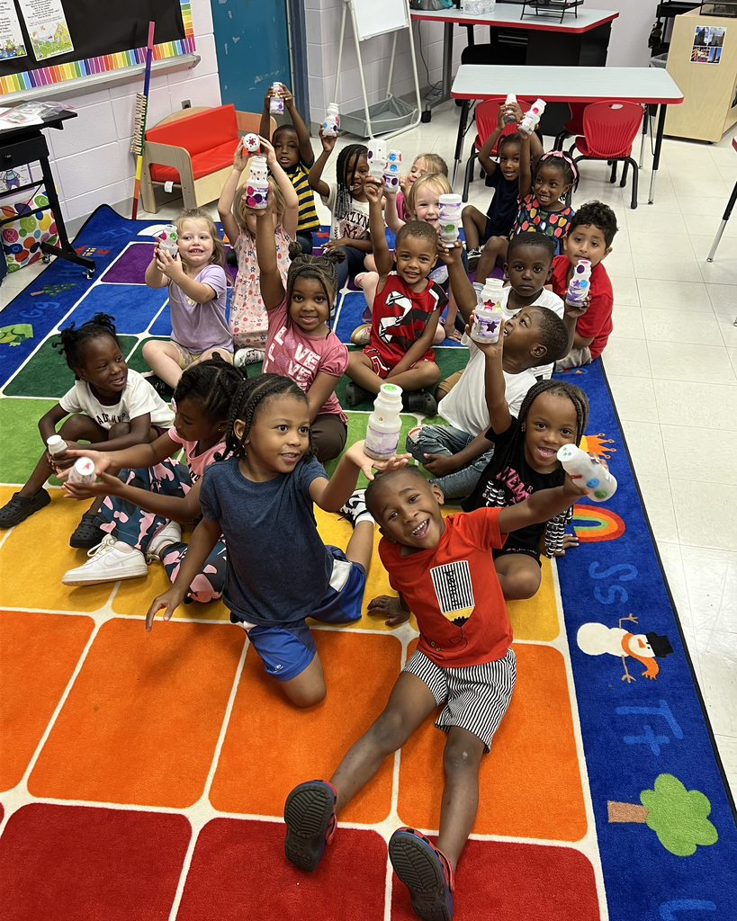 Several children sit on a colorful floor mat, holding small bottles. Some have name tags.
