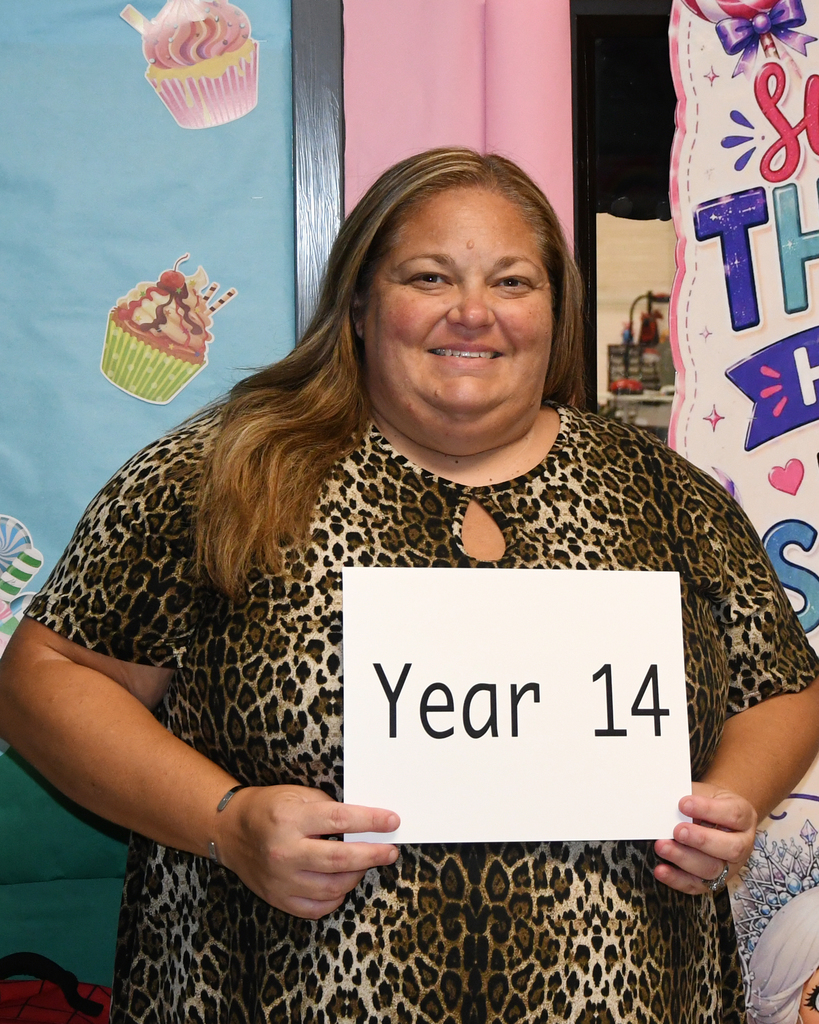 A woman in a leopard-print dress holds a sign saying "Year 14" against a colorful backdrop with cupcakes and text.