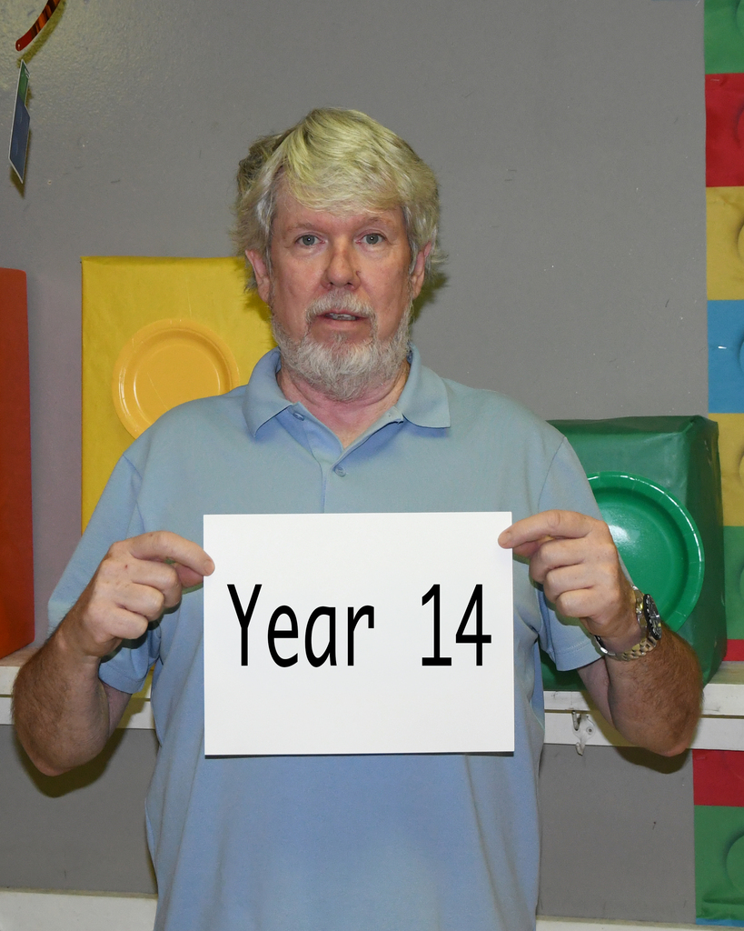 Person in light blue shirt holding a sign saying "Year 14" in front of a wall with colorful blocks.