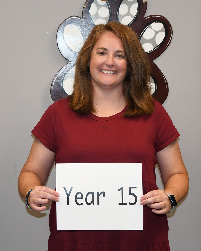 A woman with brown hair holds a sign that says "Year 15" in front of a gray background with butterfly decorations.