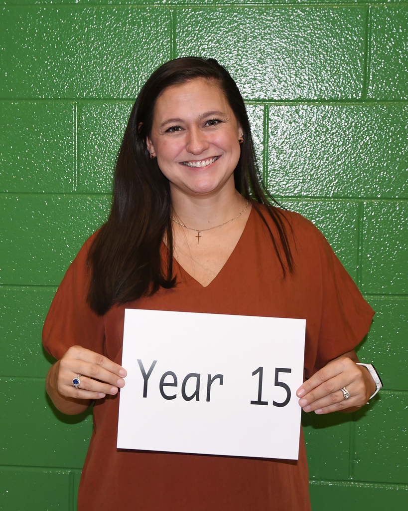 A smiling person holds a sign that reads "Year 15" in front of a green brick wall.