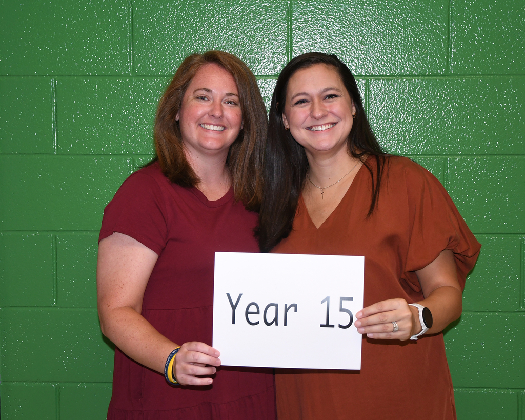Two women smiling, holding a sign that reads "Year 15" against a green brick wall.