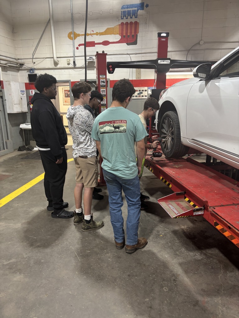 People observe a car on a lift at a repair shop. One person is working on the vehicle.