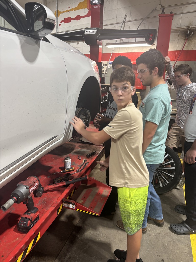 A boy stands on a jack lift, examining a vehicle tire while several others watch.