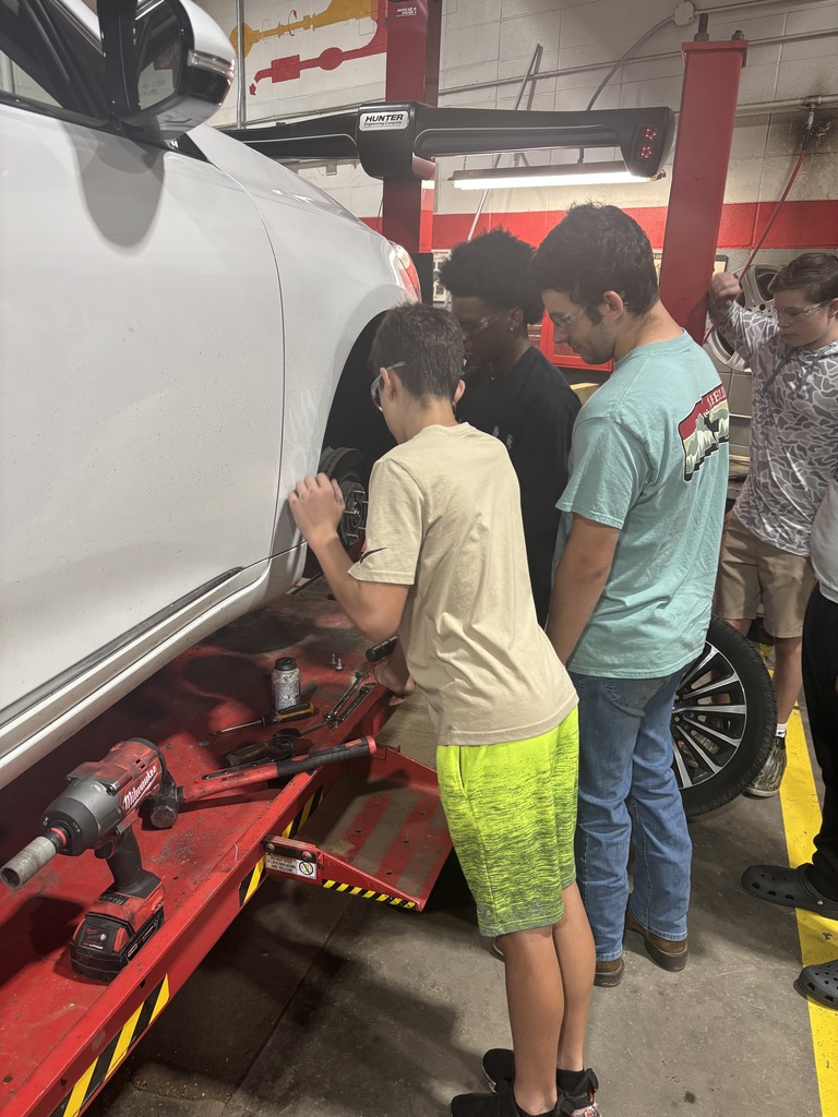 Five people in a workshop, one person is working on a white car while others watch.