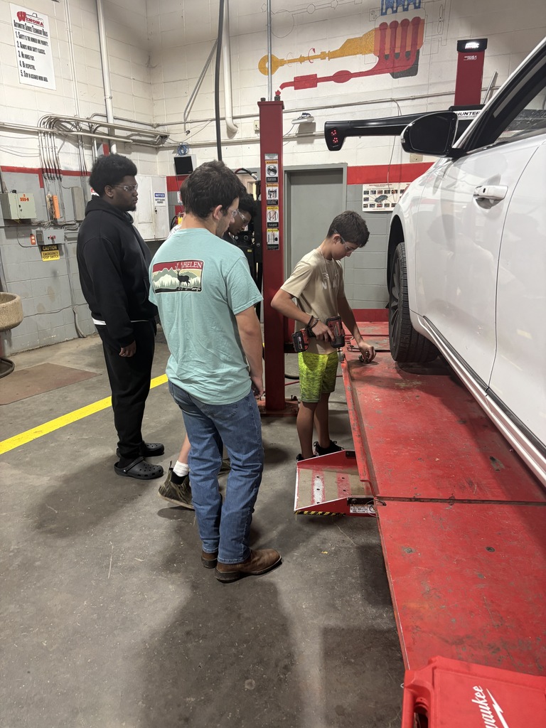 Four people in a garage are looking at a car on a red lift. One boy is standing on the lift.
