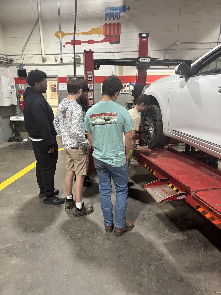 People watch as a mechanic works on a car raised on a lift inside a garage.