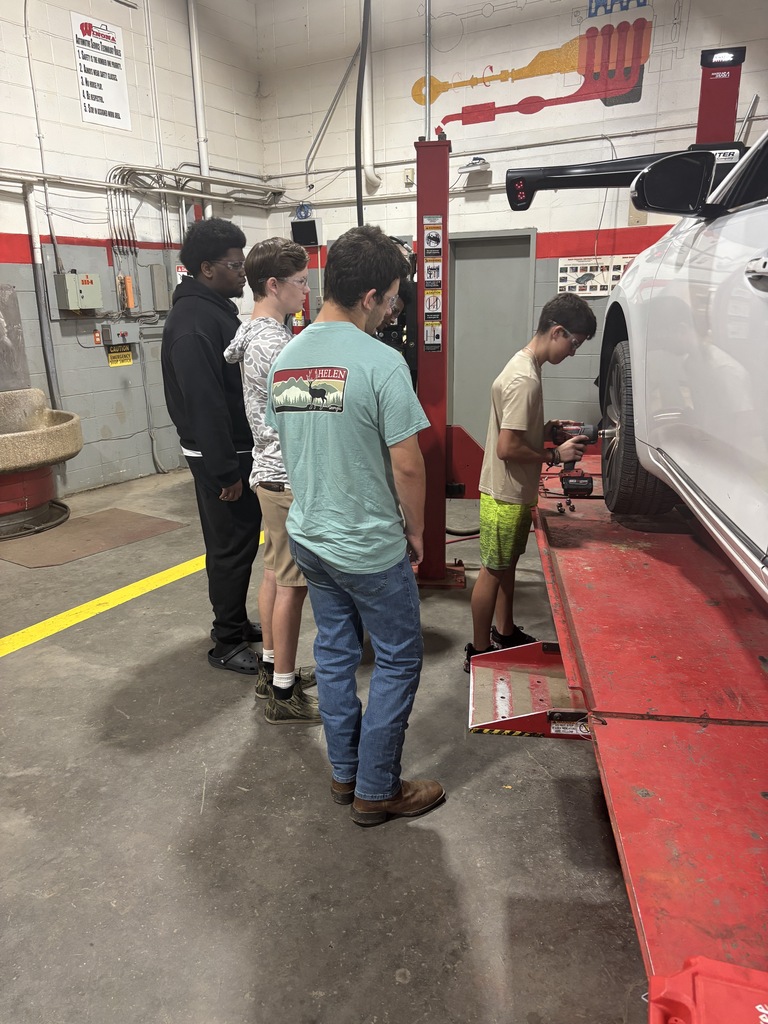 Five people, including two on jacking, work on a car in a garage. One individual bends down, and the vehicle rests on a red platform.