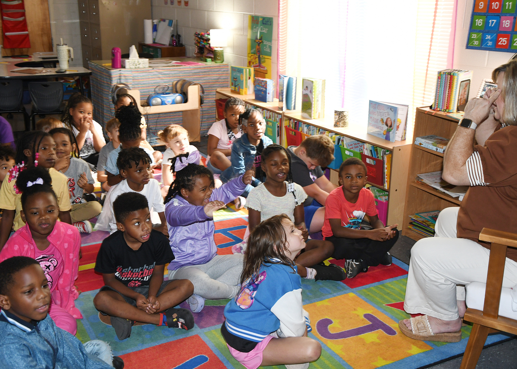 Children sitting on the floor around an adult, in a classroom with bookshelves and a rug.