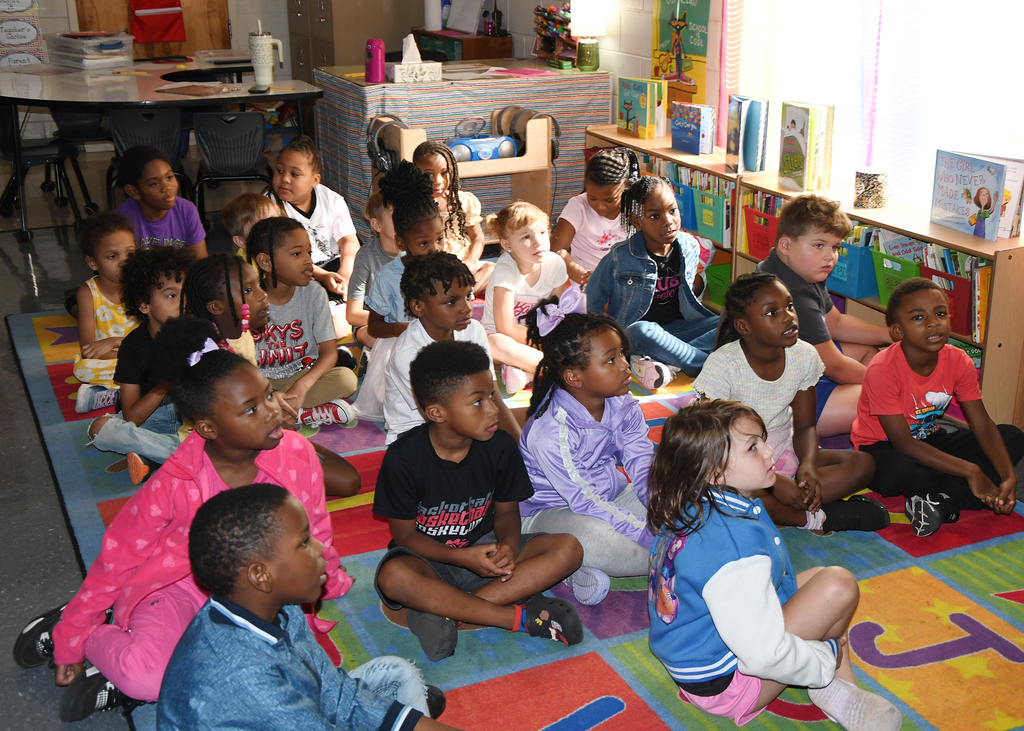Children of varying ages gather on a colorful rug inside a classroom, listening attentively to a speaker.