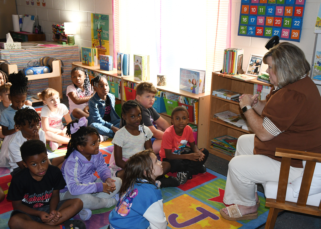 A group of children sit on a colorful rug in a classroom. An adult sits on a chair reading to them.