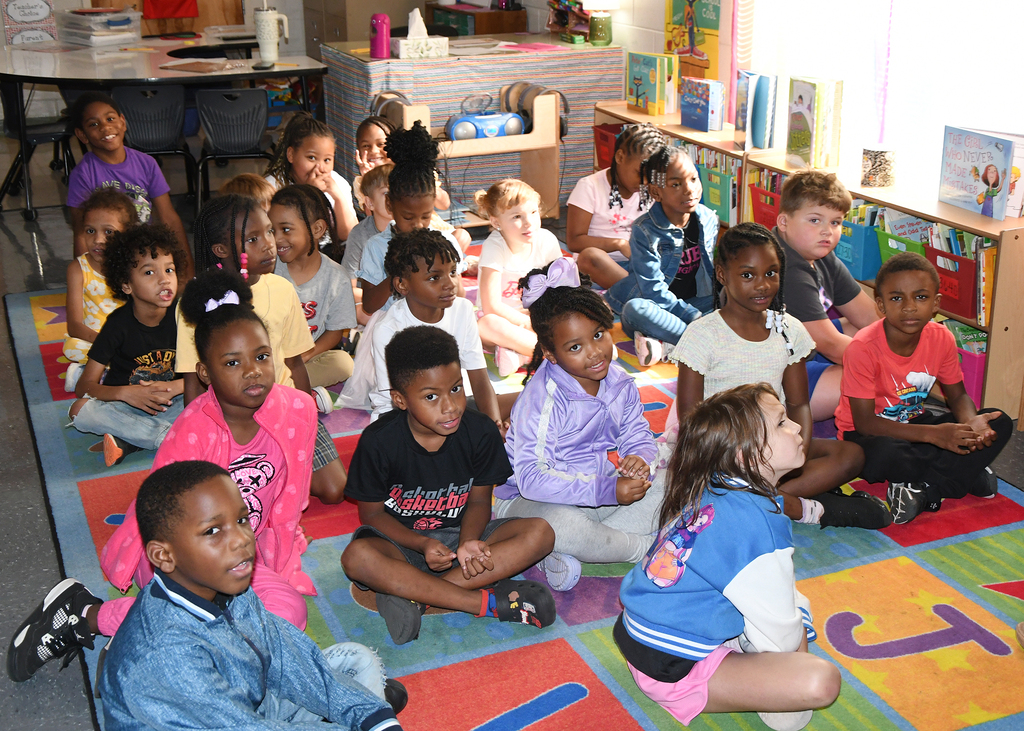 Group of children sit on a rug in a classroom, with some focused on the front and others on their surroundings.