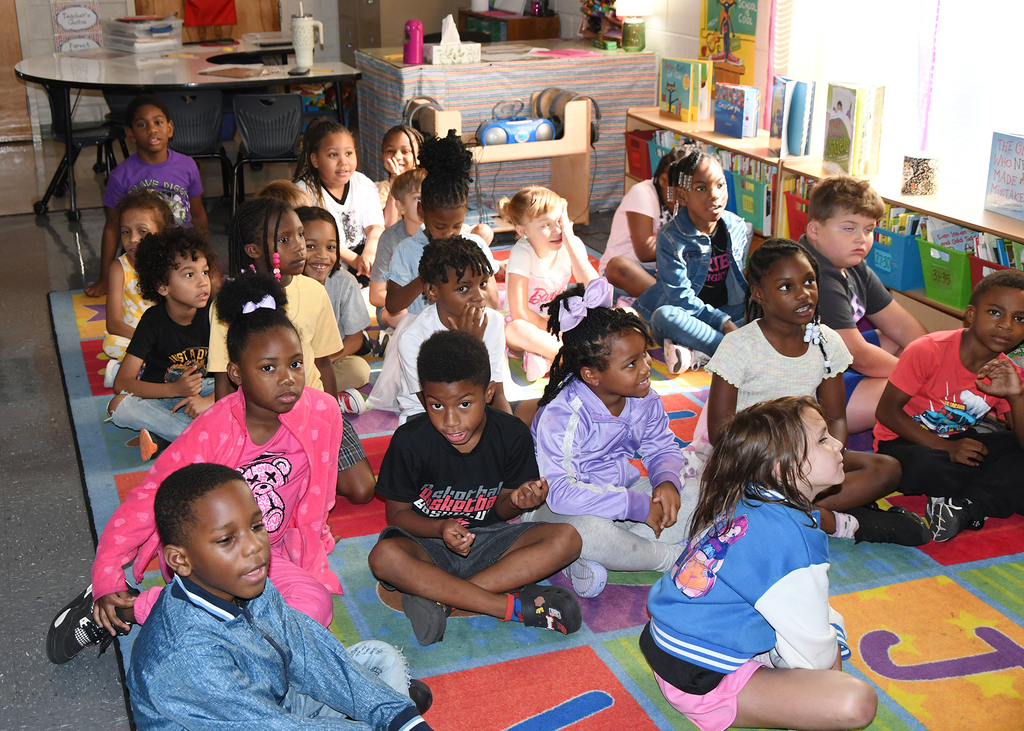 A classroom filled with children seated on a colorful rug, some in jackets, attentively listening to a teacher.