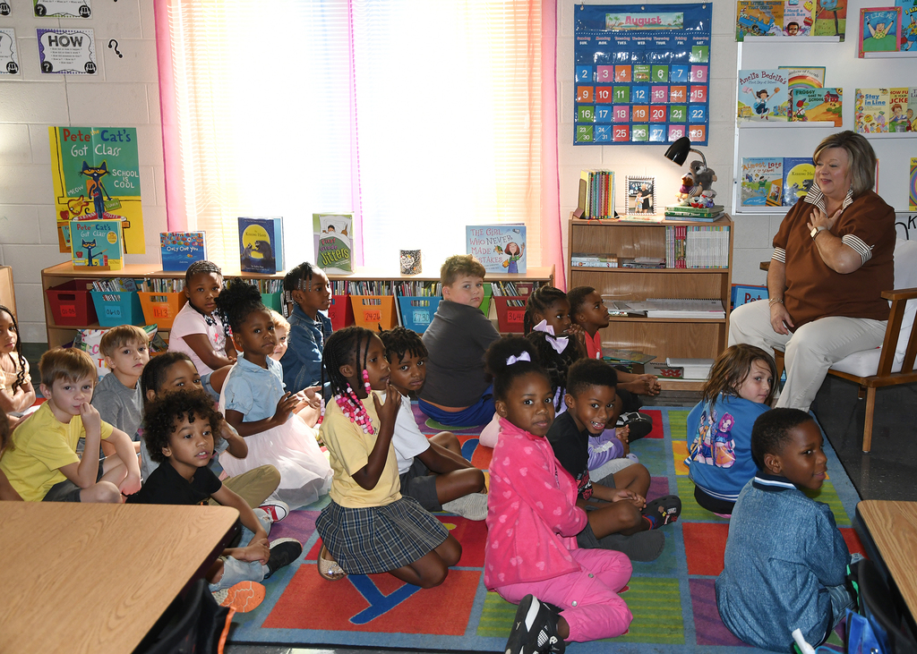 Classroom with children seated on a colorful rug listening to a teacher in front of shelves with books.