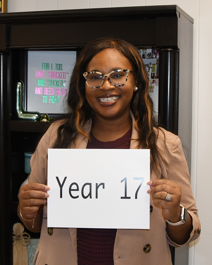 A person in a room holds a sign saying "Year 17". They have glasses, a coat, and a ring.