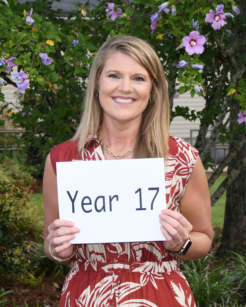 Woman in red floral dress smiling and holding a white sign that says "Year 17". Behind her is a tree with purple flowers.