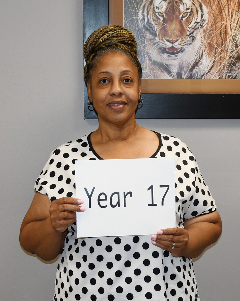 A woman in a polka-dot shirt holds a white sign that reads "Year 17" in front of her. Behind her, a tiger painting is framed on the wall.