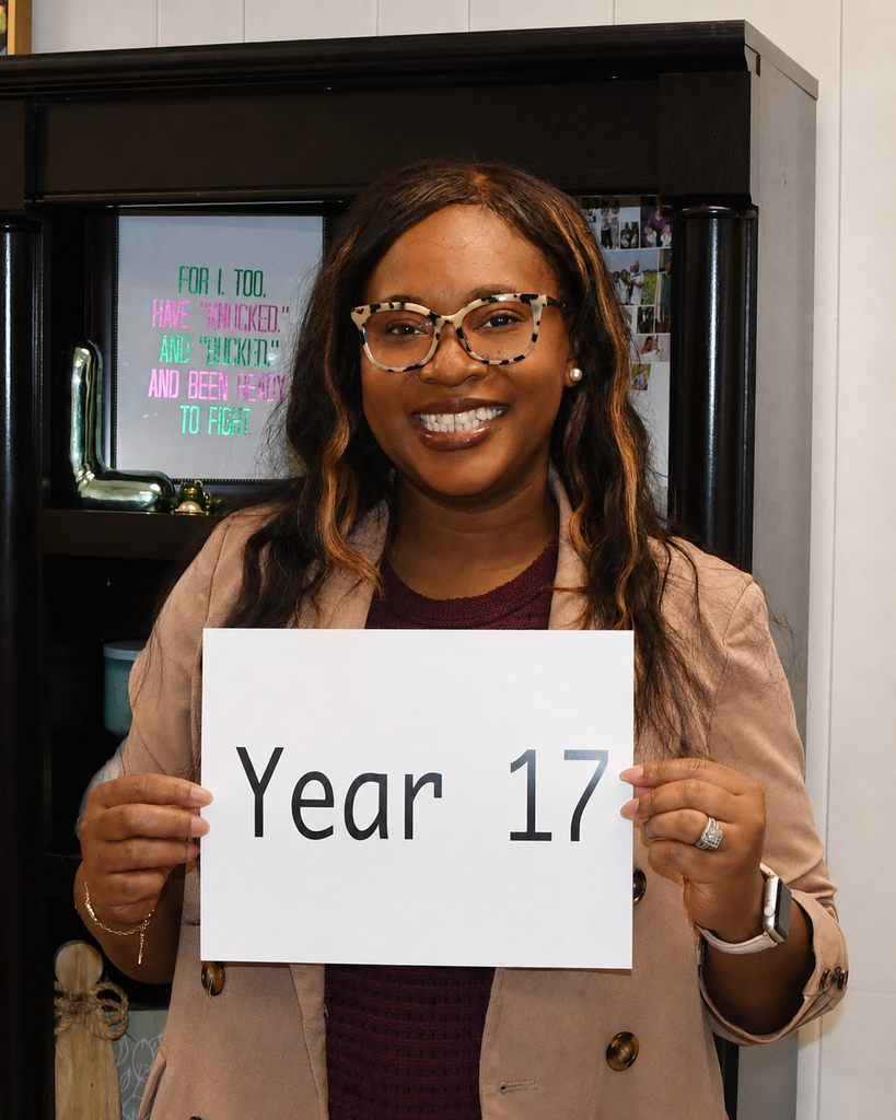 Person holding a sign that says "Year 17" in front of a bookshelf with various items.