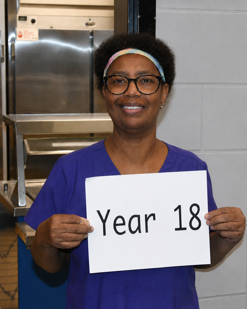 A smiling adult woman in a blue shirt holds a sign with the words "Year 18" in a kitchen area.