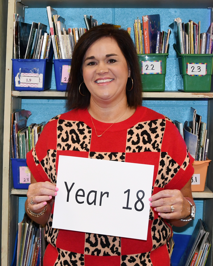 Woman in red and black patterned clothing holds a sign that reads "Year 18" in front of shelves filled with books and bins.