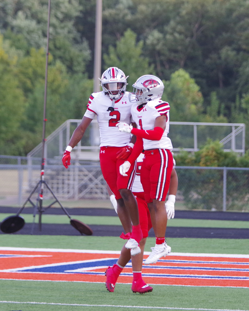 Two football players in red uniforms and helmets celebrate together on a field with a logo.