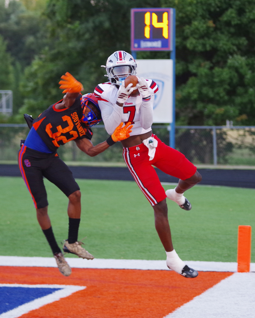 Two players, one in red and white and the other in black and blue, engage in a tackle on a field.