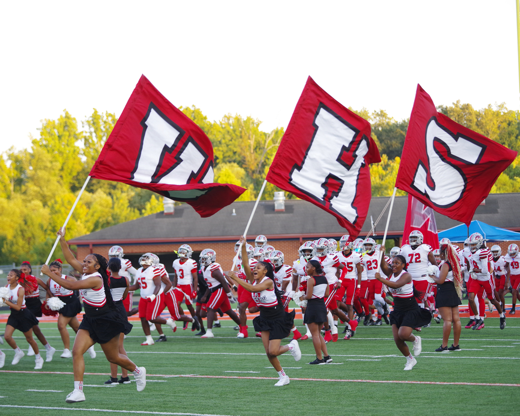 Cheerleaders wave flags with letters HS as football players run on the field. Behind them, a building has a gray roof.
