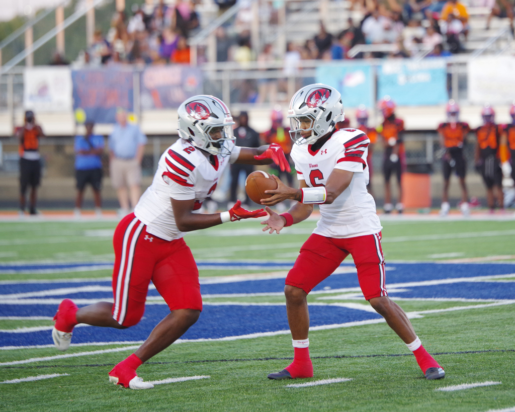 Two football players in white jerseys, red shorts, and helmets run on a field with blue stripes.