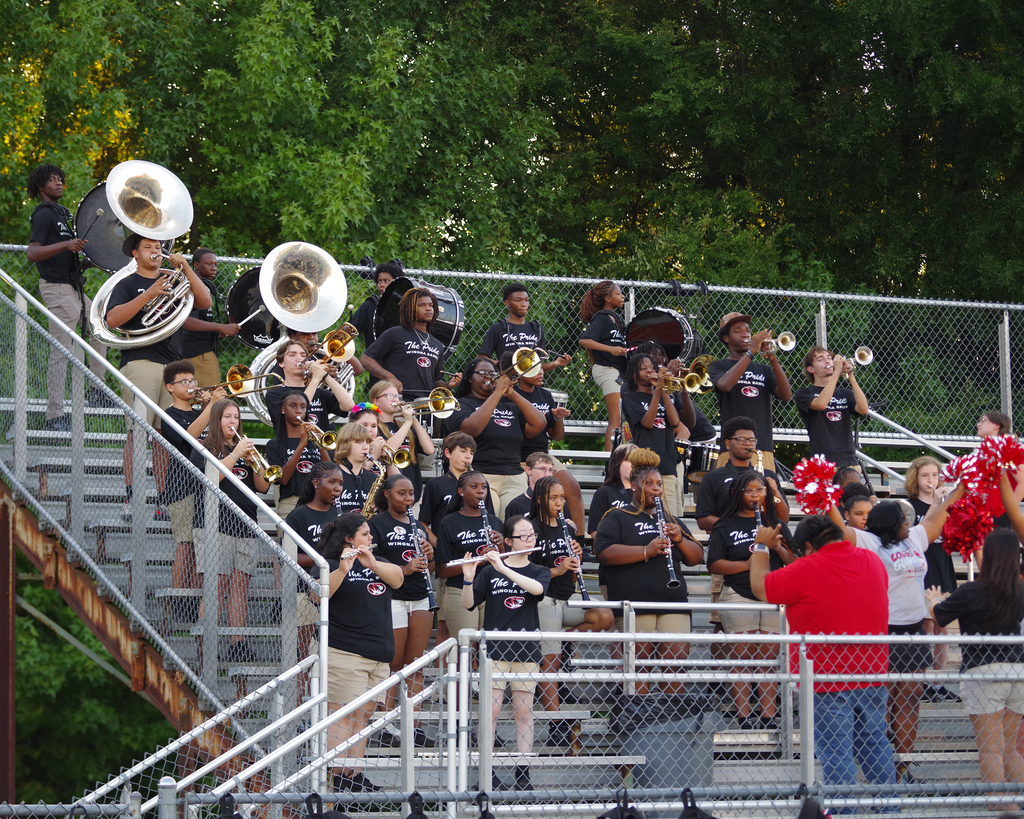 A school band stands on stadium steps, playing various instruments. Spectators in various attire watch. Trees in the background.
