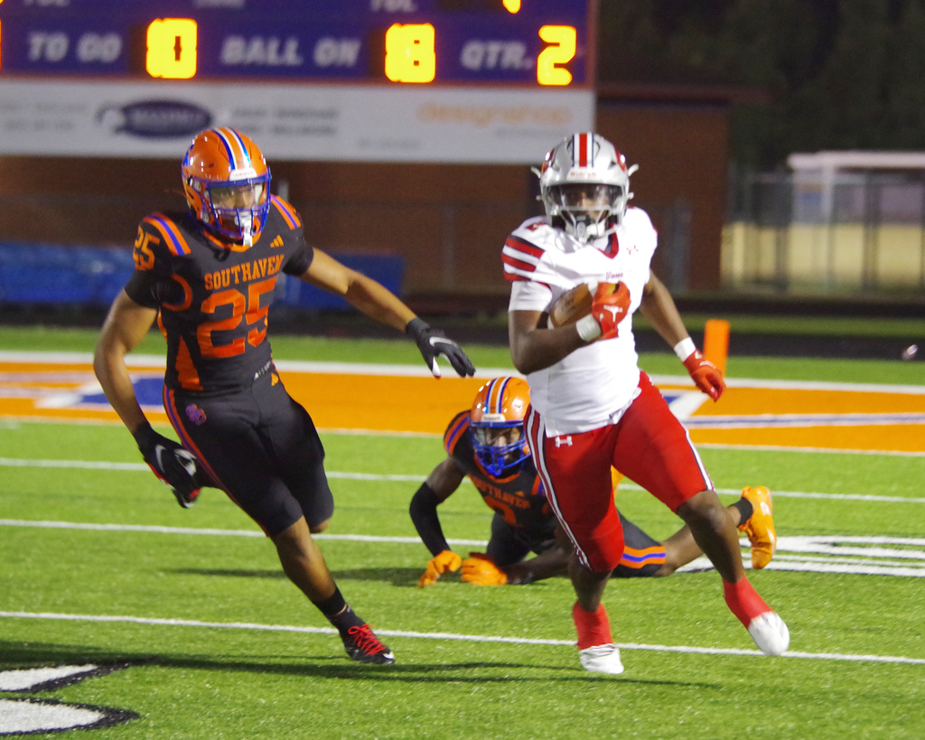 Three football players in full gear on a field. The player in white is running with the ball. The scoreboard behind them shows 0-8.