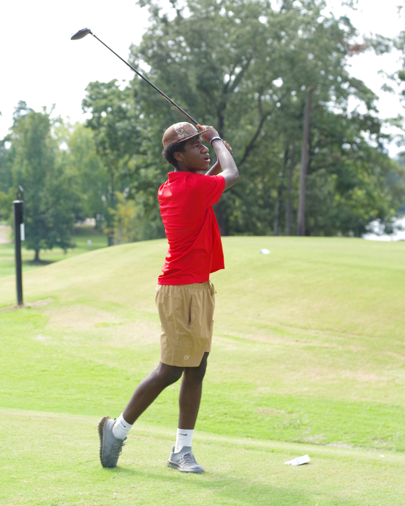 A person swings a golf club on a course. They wear a red shirt, tan shorts, and a hat. Trees and a hill are in the background.