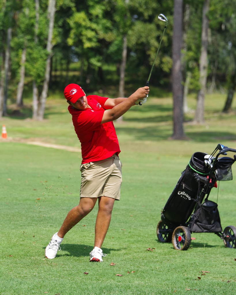A person in red shirt, shorts, and hat swings a golf club at a golf course. A golf bag is behind them.