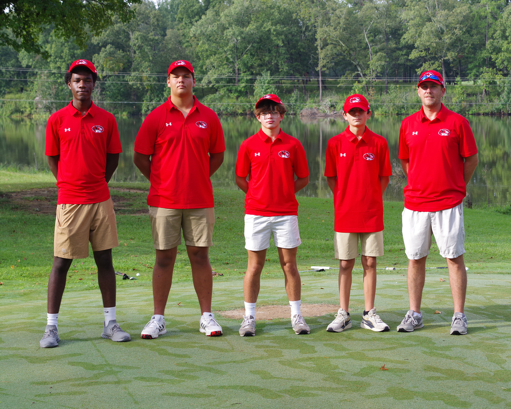 Five men in red shirts and caps stand on a golf course, facing the camera. They wear different colored shorts.