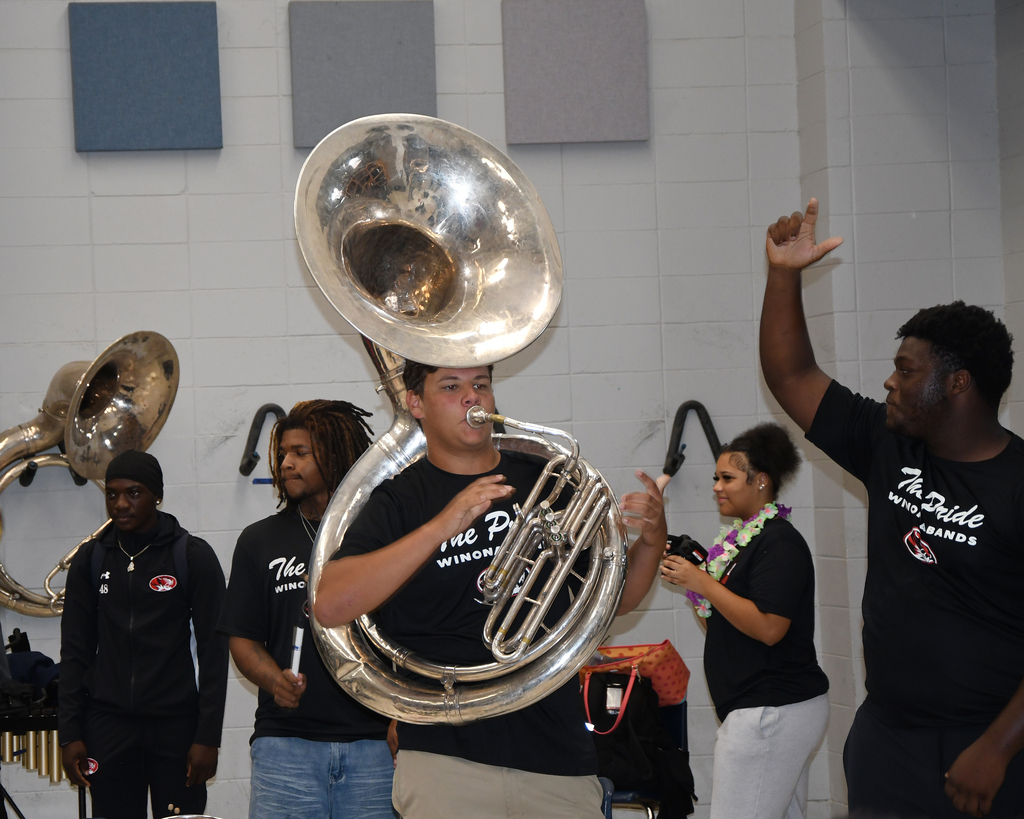 A man plays a tuba while others watch. A white wall with square patterns is in the background.