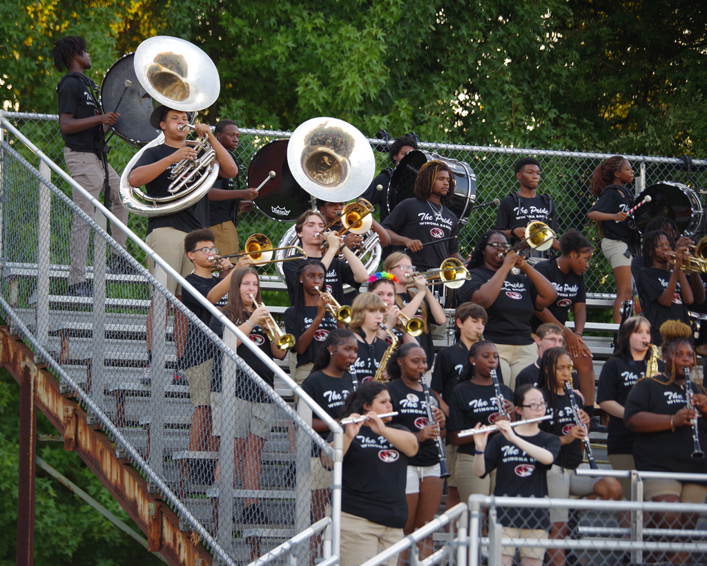 A high school band practices on bleachers with instruments, including tubas, trumpets, and flutes, on a sunny day.