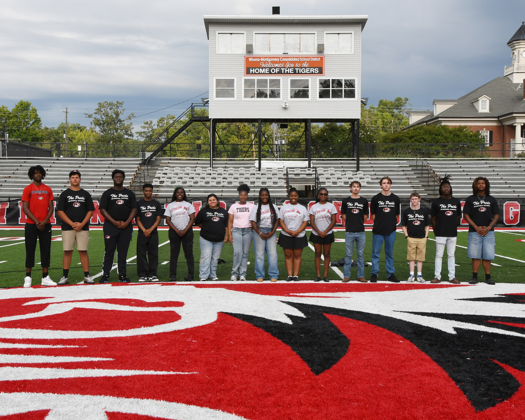 A group of 13 individuals, dressed in black, stand on a football field. Behind them are bleachers and a structure.