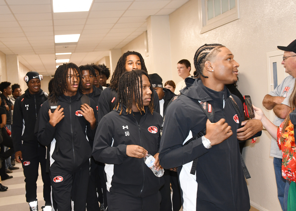 A group of athletes wearing matching black jackets walk through a hallway. They are approached by others.