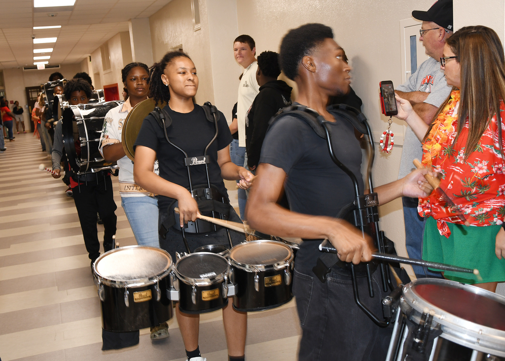In a school hallway, several students line up, playing drums with harnesses on their bodies.