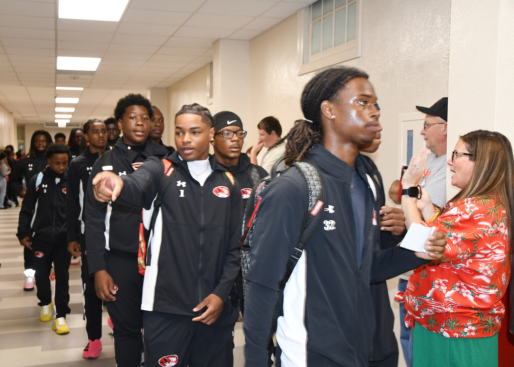 Athletes in matching black uniforms walk in a hallway. A woman in a red floral shirt speaks to one of them.
