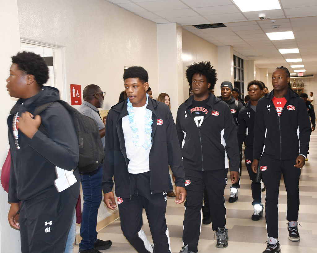 A group of people wearing matching black jackets and pants walk down a corridor. Some have white undershirts.