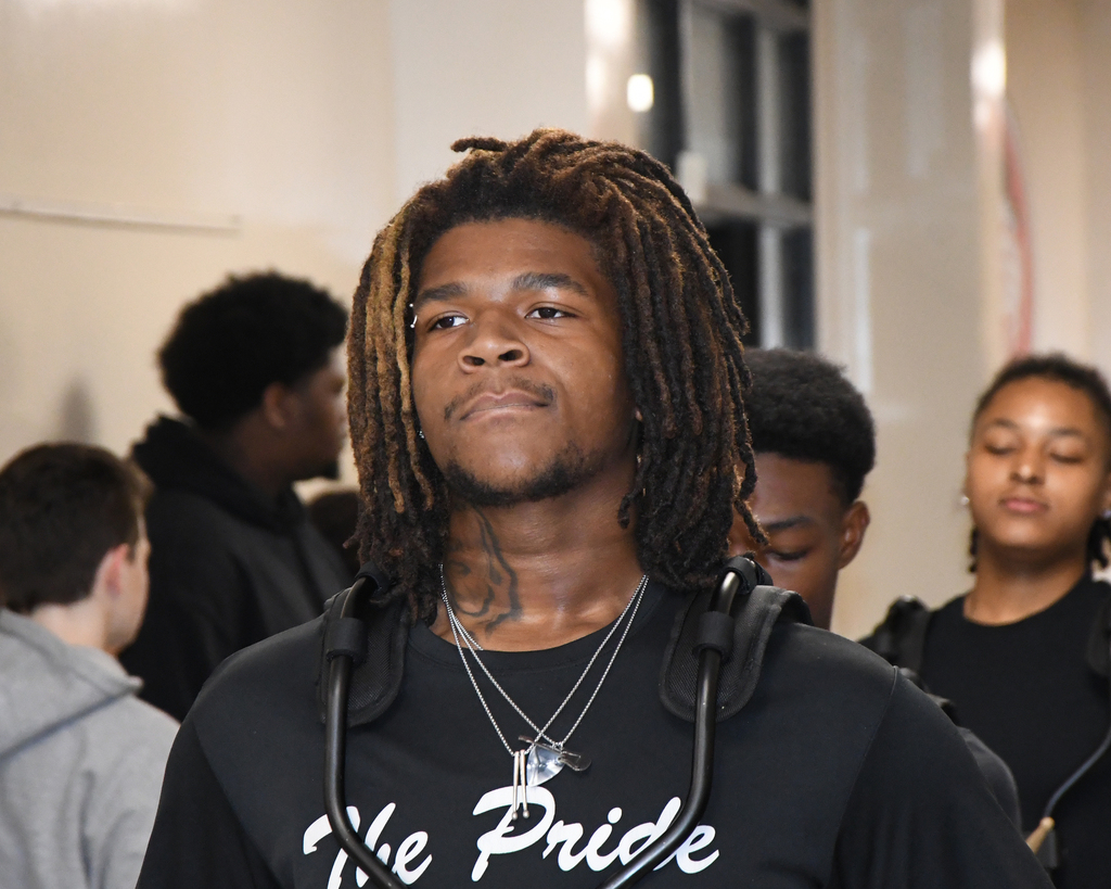 A man with dreadlocks and a black shirt reading "The Pride" stands with others in a hallway.