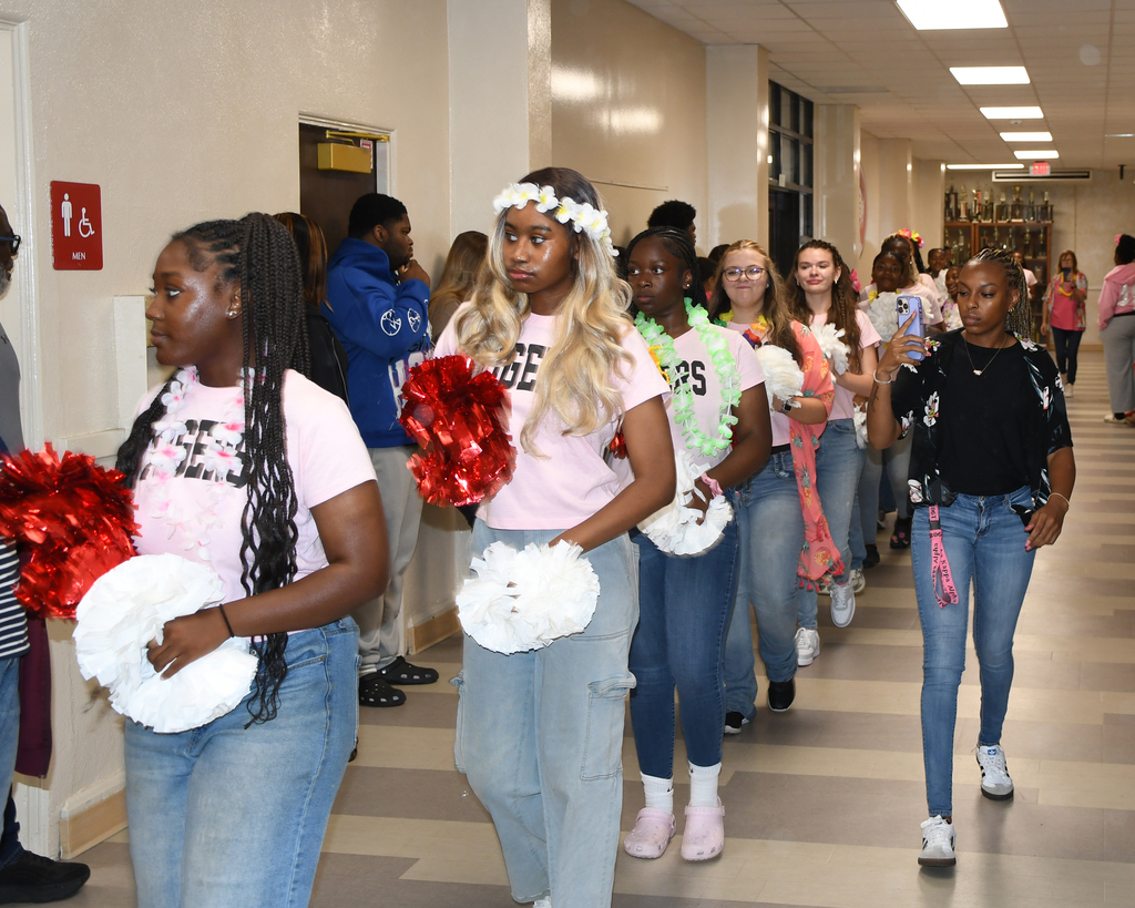 A group of young people walking down a hallway. They wear pink shirts, jeans, and hold pom-poms.