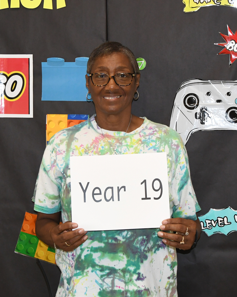 A woman holds a white sign that reads "Year 19" in front of a backdrop featuring gaming and building block graphics.