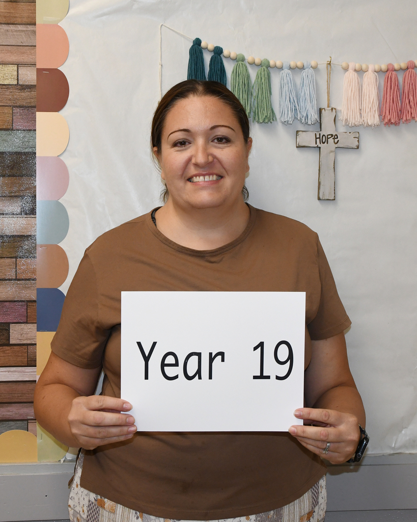 A smiling woman holding a sign that says "Year 19" stands against a backdrop of multicolored wall tiles and a cross.