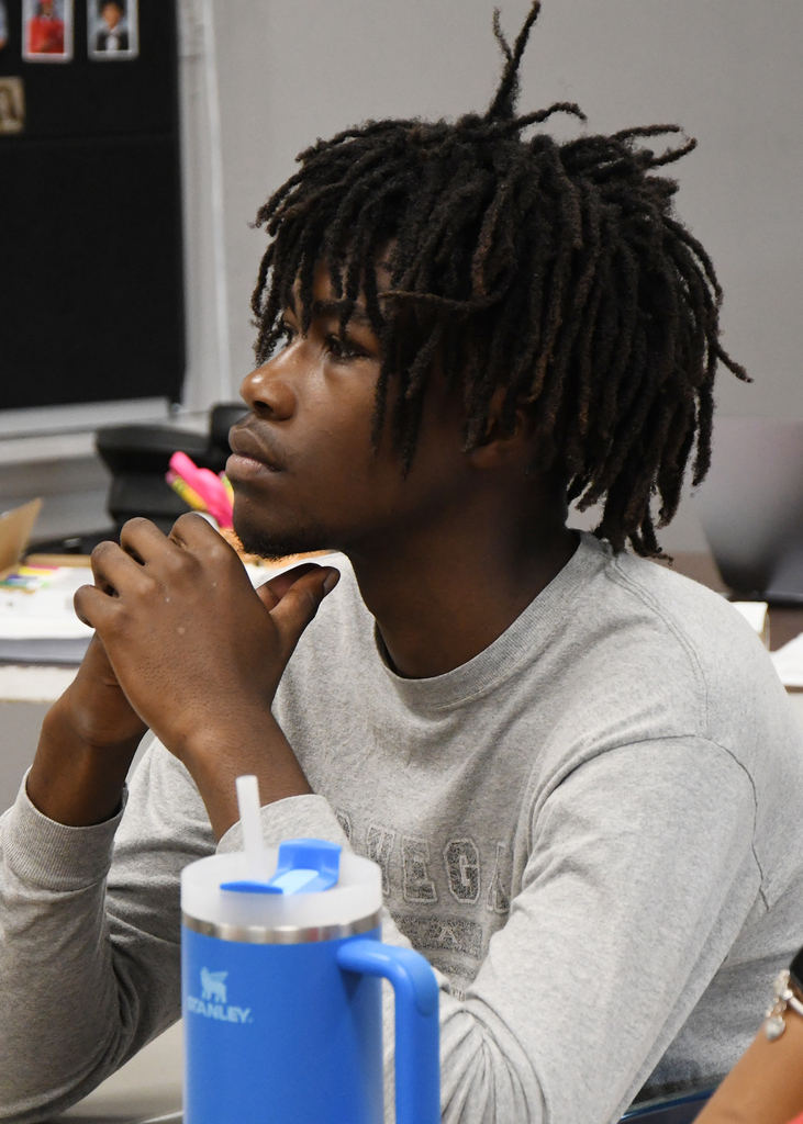 Person with dreadlocks in a gray shirt sits in a classroom. A blue tumbler sits on the table.