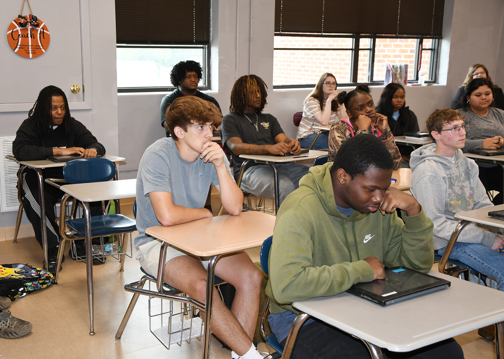 Students seated in a classroom, some with laptops, appear focused on their tasks. Windows with blinds and a basketball poster are visible.