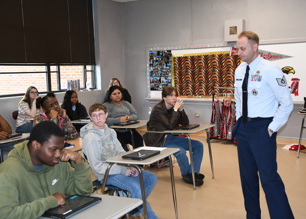 A man in uniform stands before students seated at desks in a classroom with windows and decorations.