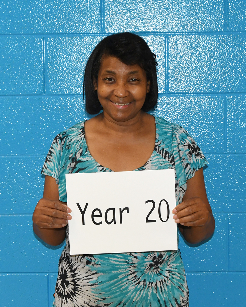 Person holds a white sign reading "Year 20" against a blue brick wall, smiling and posing for a photo.